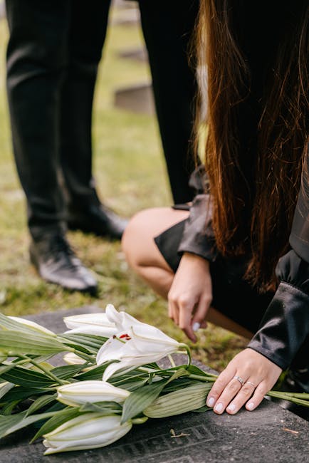 A person with long brown hair wearing a black dress is kneeling on the ground, adjusting a carefully arranged bouquet of white lilies with large green leaves, wrapped in clear cellophane. The lilies a