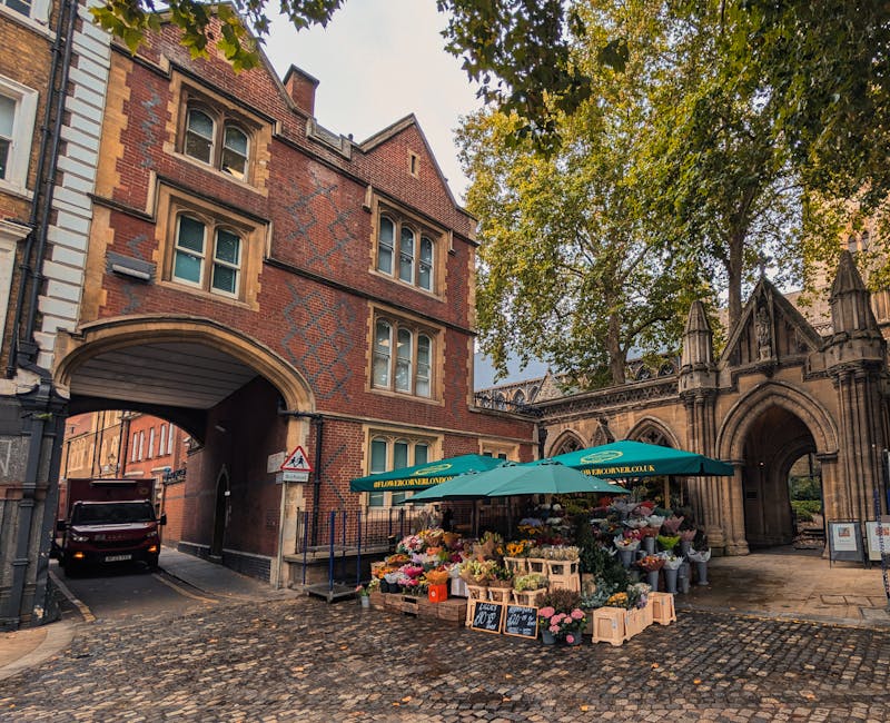 A vibrant outdoor flower stall situated on a cobblestone street in front of a historic brick building with arched entryways, featuring a variety of fresh flowers including roses, lilies, and daisies i