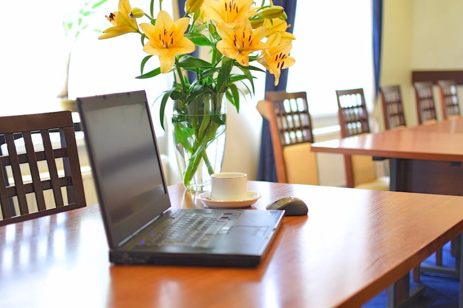 A clear glass vase filled with vibrant yellow lilies and long green stems sits atop a polished wooden table in a well-lit conference room. The arrangement showcases the large, trumpet-shaped petals of