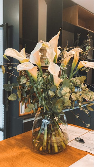 A floral arrangement featuring elegant ivory calla lilies with smooth, elongated petals and dark green, rounded eucalyptus leaves, arranged in a clear glass vase filled with water. The bouquet is plac