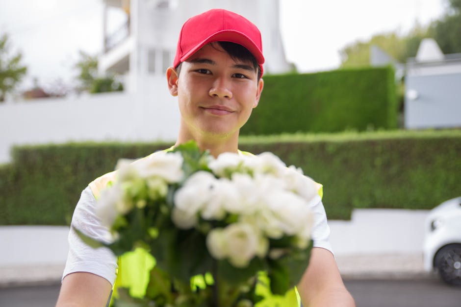 A delivery person wearing a red cap and high-visibility vest is holding a bouquet of fresh white roses and green foliage, extending it towards the camera in front of a residential area with white hous