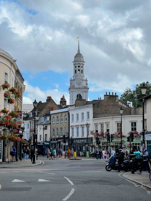 The image shows a busy street scene in Balham with a prominent Victorian clock tower in the background, under a partly cloudy sky. In the foreground, there are storefronts decorated with hanging flowe